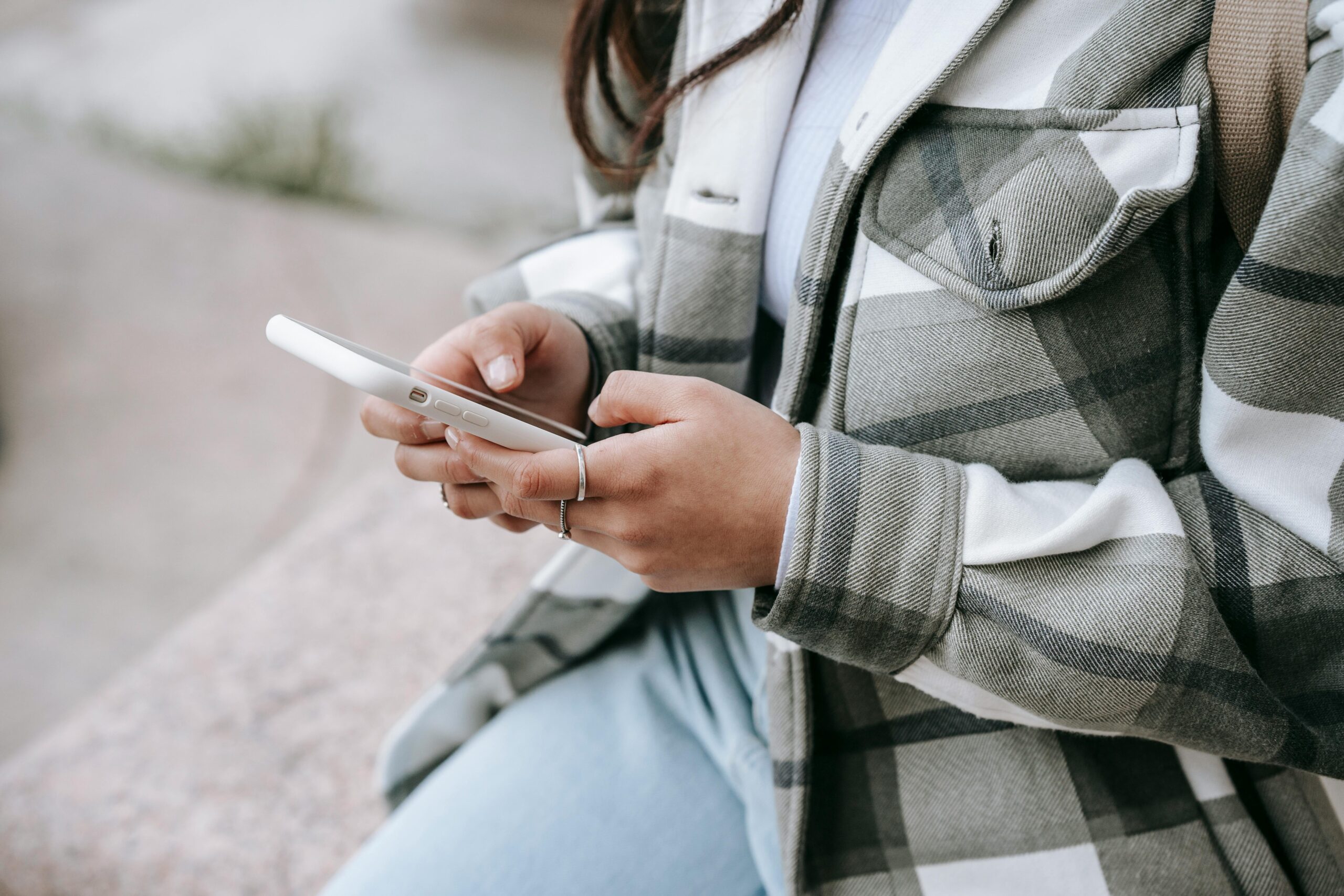 anonymous-young-woman-in-casual-outfit-sitting-on-stone-fence-and-text-messaging-on-phone-in-daytime